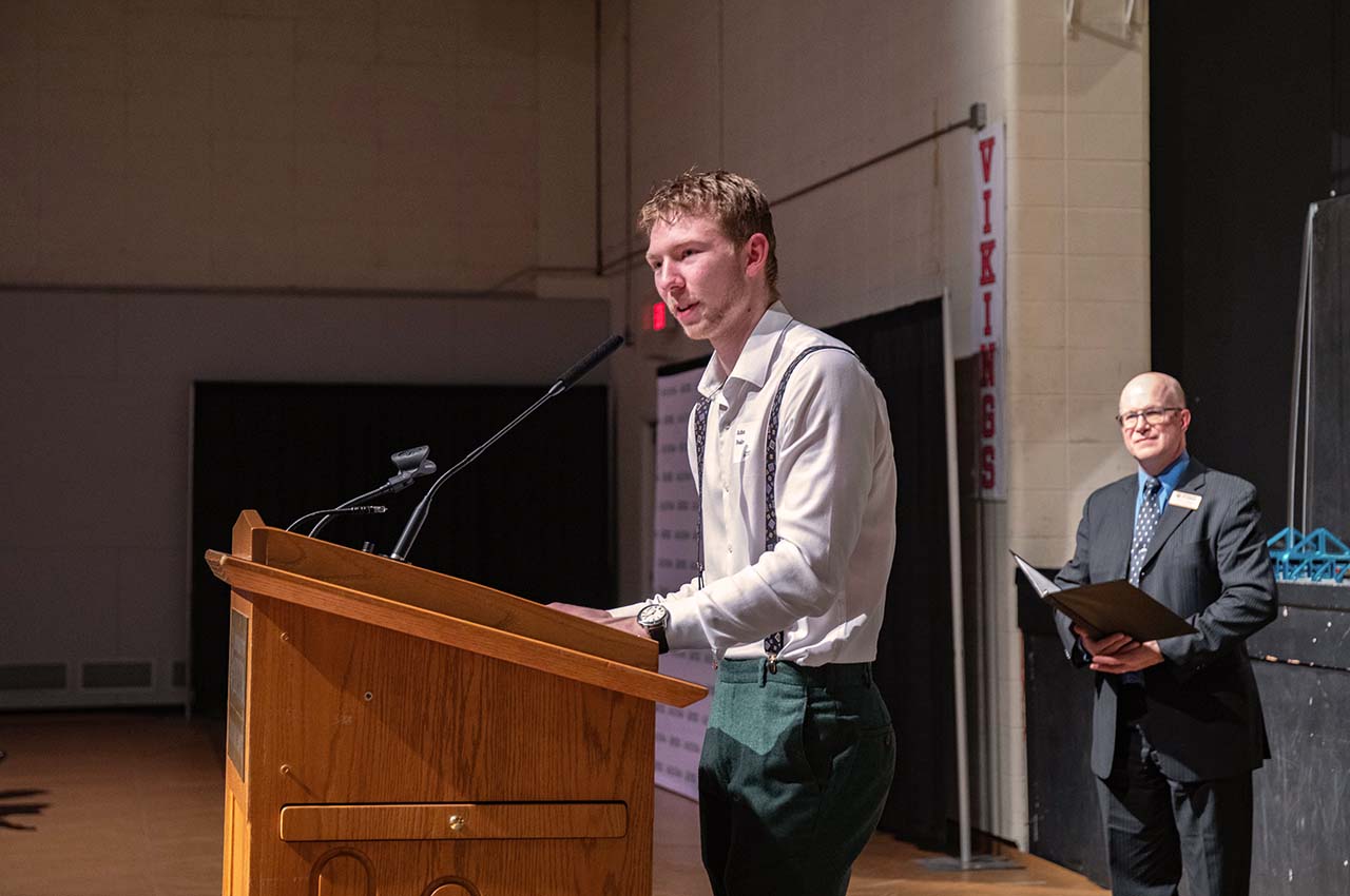 A student peaking at a podium, a staff member standing in the background.