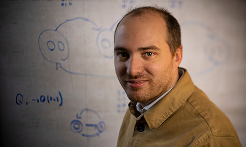 A 3/4 profile of Thibaud Lutellier looking at the camera and smiling in front of a white board with writing on it.