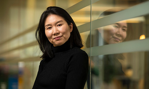 Photo of Mi-Young Kim wearing a black turtleneck as she leans against a glass wall as she smiles at the camera, her reflection seen in the glass.