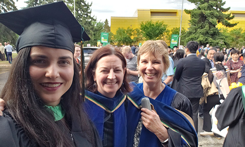 Three women wearing academic robes taking a selfie outside the Northern Jubilee Auditorium on North Campus. 