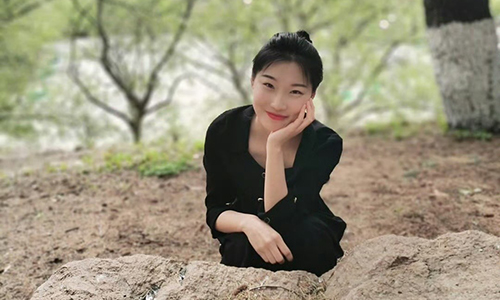 Fengjing An crouching down and smiling at the camera in a wooded area. 