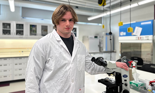 Daimen Asp in an Augustana Biology lab, wearing a lab coat and hand on a microscope.