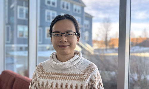 Huanqing Chen standing in the Augustana Forum building, Founders' Hall viewable through the window behind her.