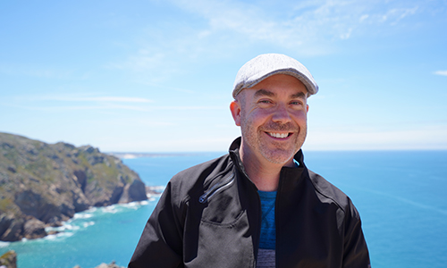 Scot Lorenson wearing a page boy hat and smiling in front of a view cliff side and the ocean.