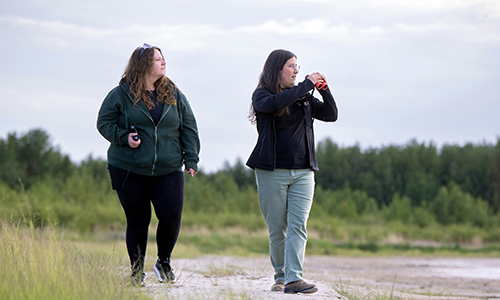 Two people walking outdoors and looking off into the distance, one of them holding a pair of binoculars.