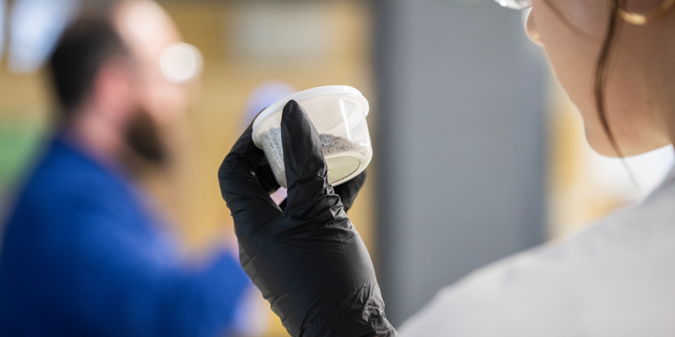 Woman holding a plastic container with dirt in it. Wearing a black rubber glove and while lab coat