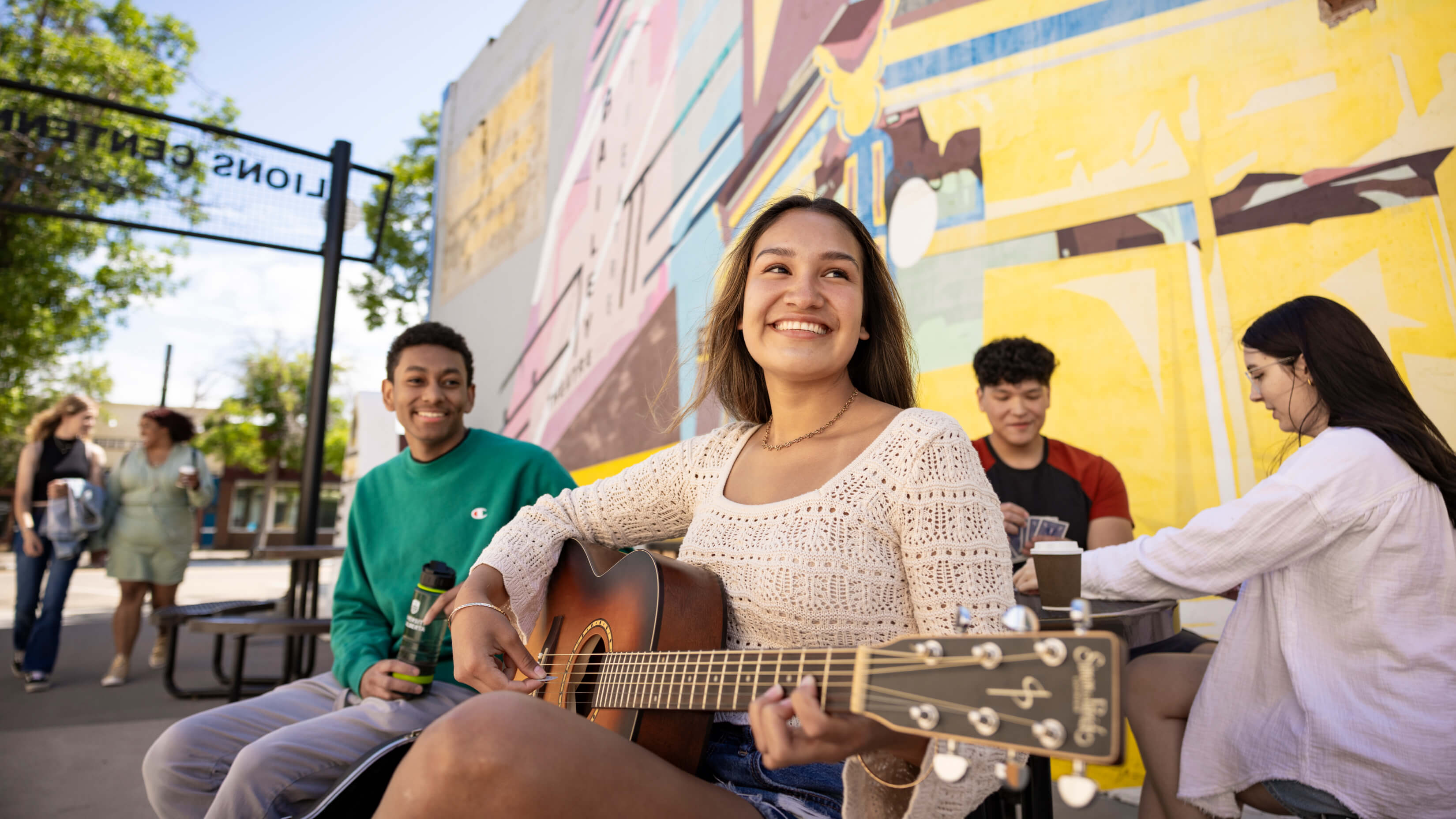 Smiling female student playing acoustic guitar outdoors with friends gathered around
