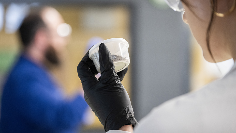 Student wearing lab PPE while examining container of granular material in an Augustana lab.