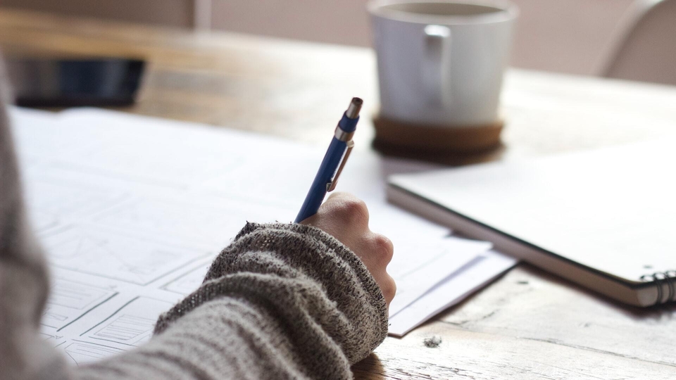 A student works at a table with a pen and paper with a mug of a warm drink in the background.