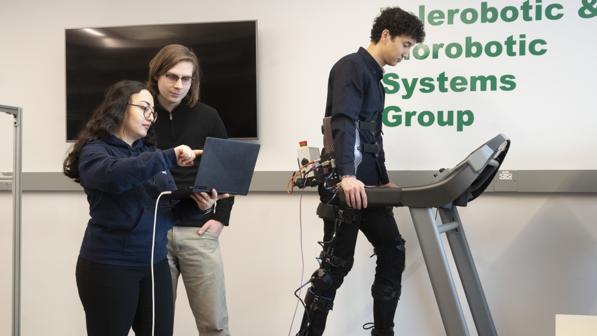 A man on a treadmill wearing robotic gear, another man and woman staring at a laptop screen