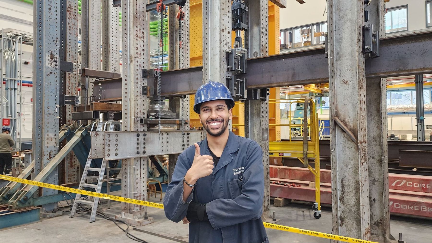 Smiling man in a hard hat holding a thumbs up in a factory