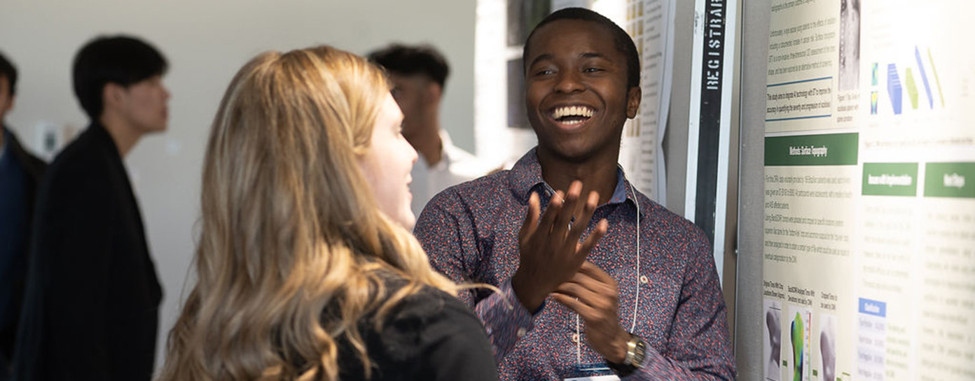 A student laughs while taking questions about their research in front of their research poster.