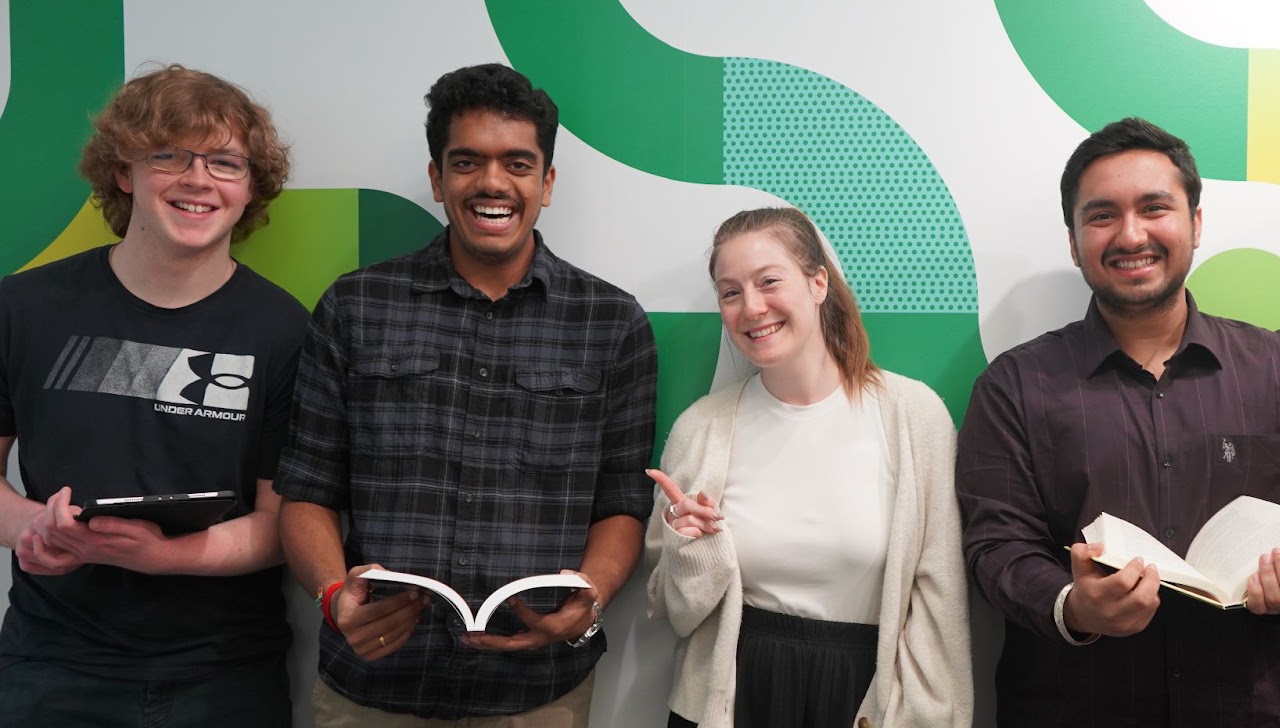 Four smiling students stand together in front of a white and green patterned wall.