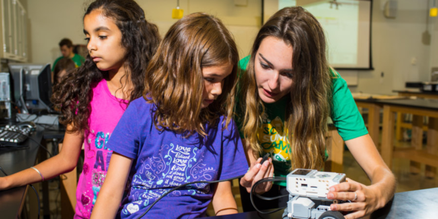 A computer science camp leader shows a child a small computer chip while another child looks at a computer off-frame
