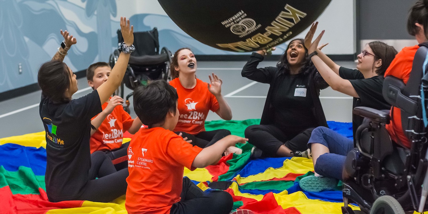 Children, teenagers, and camp leaders sitting in a circle play with a kinball (very large volleyball) in a Free2BMe summer camp