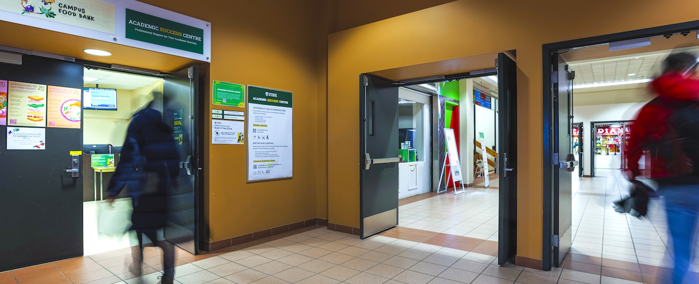 View of the entrance of the Academic Success Centre inside SUB mall with a firgure walking into it, and another figure walking through the doors to the rest of the mall