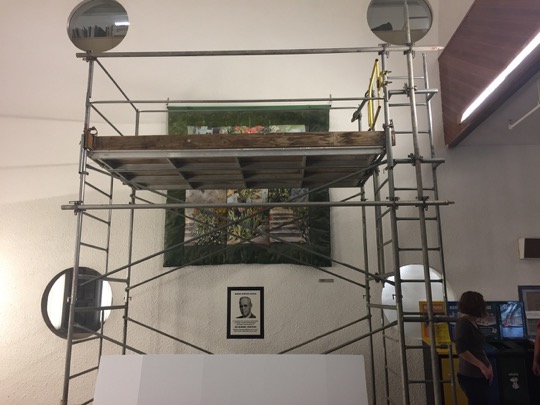 A large work of art hangs on a wall surrounded by scaffolding