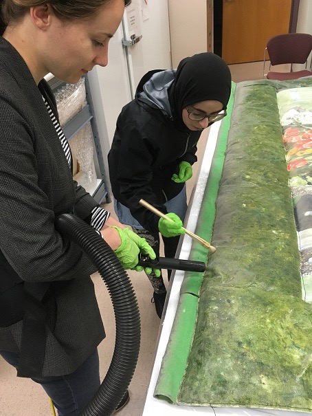 Two individuals holding cleaning tools against a large museum object