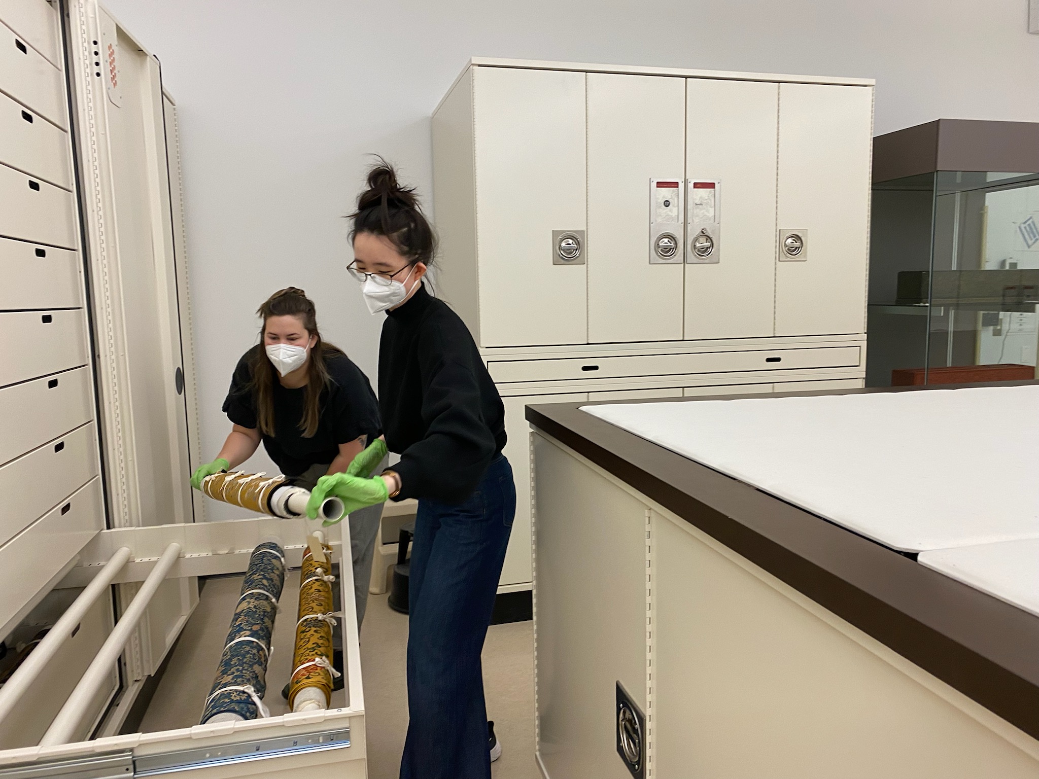Two individuals carefully place a rolled textile into a pulled out storage cabinet. 