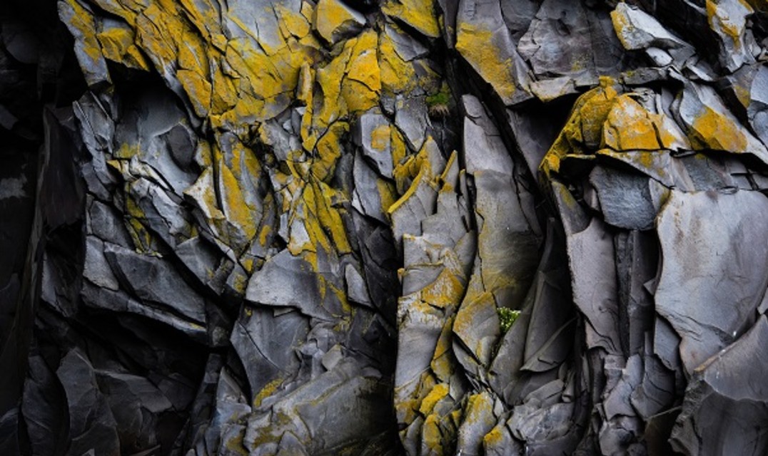 Closeup of a grey, jagged rock face with flecks of yellow discoloration and moss