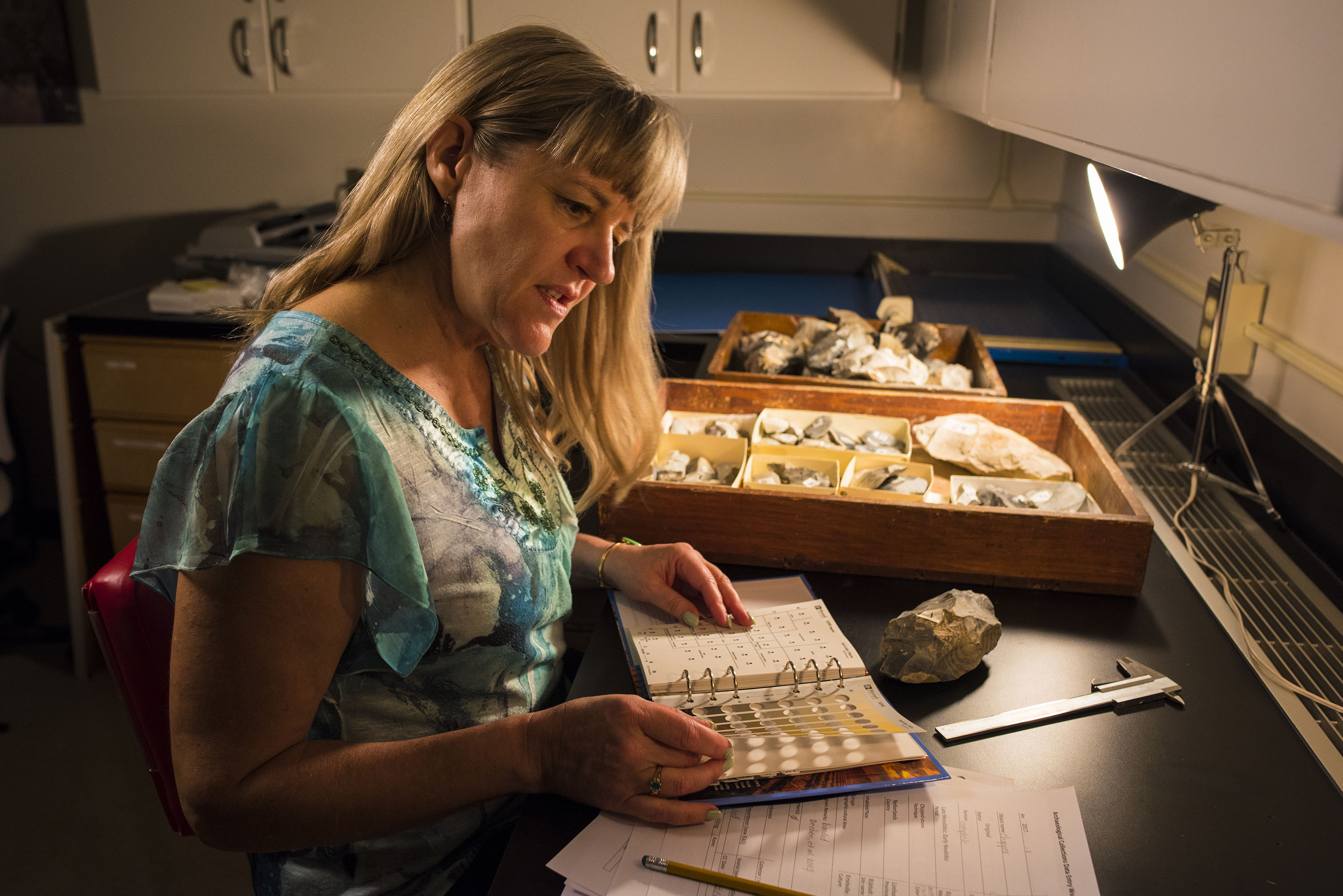 A woman sits at a desk while reading a binder of information surrounded by artifacts.